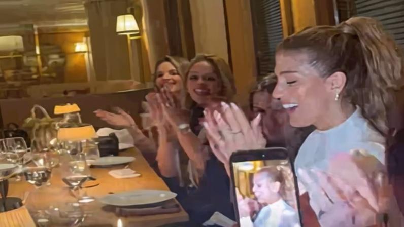 À esquerda, grupo de cinco pessoas sentadas à mesa de jantar, sorrindo e aplaudindo, com uma pessoa segurando celular mostrando vídeo chamada. À direita, mesa com taça de vinho, talheres e cardápio do restaurante L'Avenue Paris.