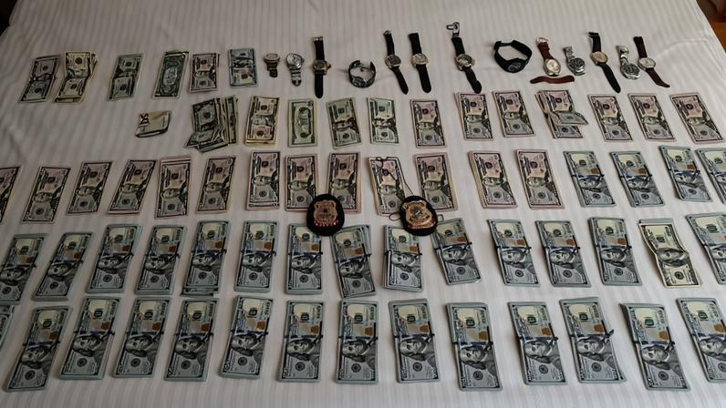 Diversas cédulas de dólares organizadas em fileiras sobre uma cama branca, acompanhadas por vários relógios de pulso e dois distintivos policiais no centro da disposição.