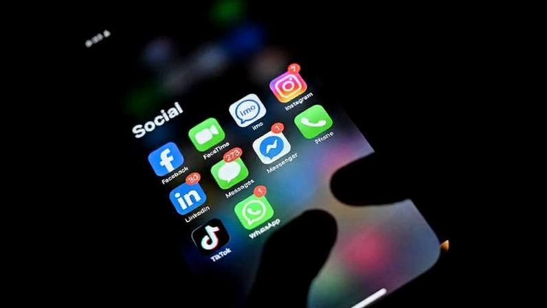 Tela de smartphone exibe pasta chamada 'Social' com ícones de Facebook, FaceTime, imo, Instagram, LinkedIn, Mensagens com 273 notificações, Messenger com 1 notificação, Telefone, TikTok, WhatsApp com 9 notificações. Fundo escuro com mão segurando o aparelho.