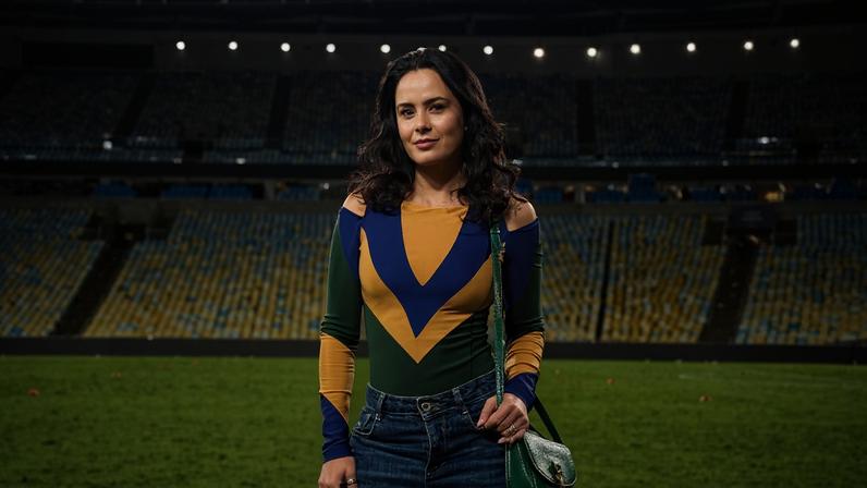 Mulher de cabelos soltos veste blusa colorida com padrão em V e calça jeans larga, posando no gramado de estádio de futebol vazio à noite, com arquibancadas e iluminação artificial ao fundo. A atriz Luciele Di Camargo: 'Também precisei colocar minha vida em segundo plano'