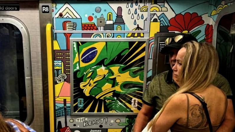 Interior de vagão de metrô decorado com arte colorida que inclui várias bandeiras do Brasil no teto. Porta aberta mostra plataforma com duas pessoas caminhando e colunas vermelhas.
