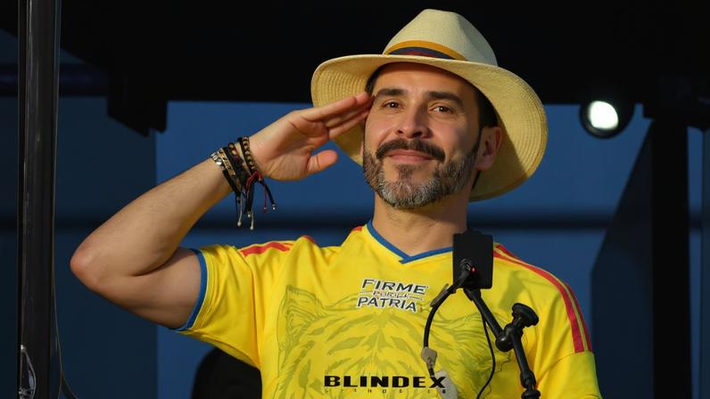 Homem de barba e bigode usa chapéu branco e camiseta amarela com inscrições, fazendo saudação com a mão direita. Ele está atrás de um vidro com microfone fixado à frente.