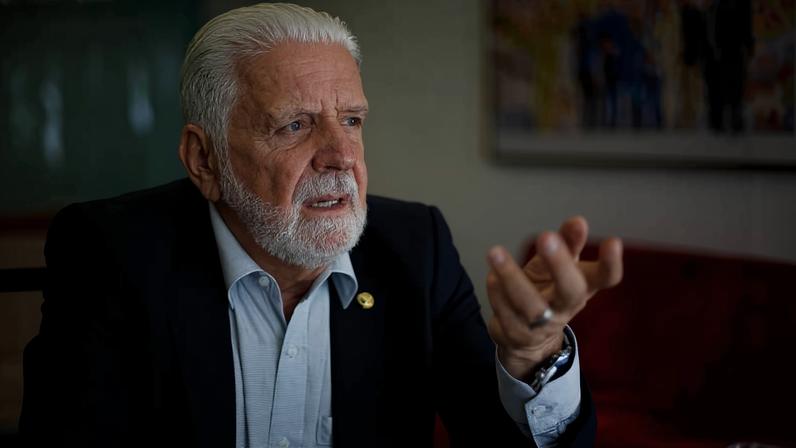 Homem branco idoso com cabelo e barba grisalhos, vestido com camisa azul clara e paletó escuro, gesticula com a mão direita enquanto fala em ambiente interno com parede clara e quadro desfocado ao fundo.