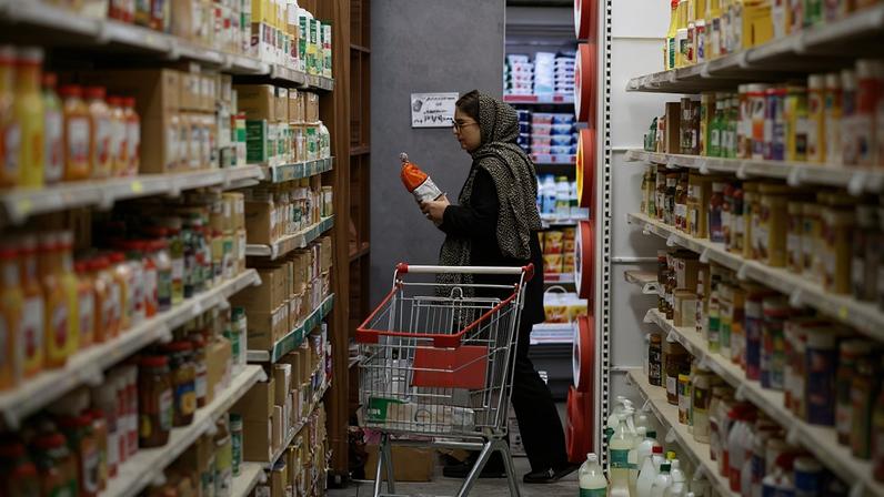 Pessoa com capuz escuro segura uma garrafa laranja enquanto está entre prateleiras cheias de produtos em corredor de supermercado. Carrinho de compras parcialmente cheio está ao lado.