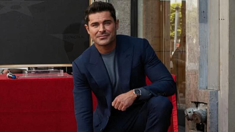 Homem vestido com terno azul escuro e camiseta cinza ajoelhado ao lado de estrela na Calçada da Fama de Hollywood, com tapete vermelho ao redor e placa da Calçada da Fama ao fundo. 'Por muito tempo, foquei apenas no trabalho e negligenciei minha vida pessoal', diz Zac Efron