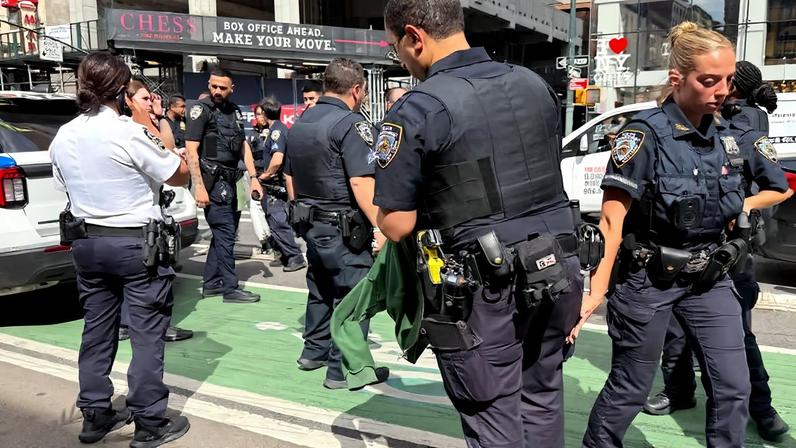 Seis policiais uniformizados estão reunidos em uma faixa de ciclovia verde em uma rua movimentada de Nova York. Ao fundo, há veículos policiais estacionados e uma placa com o texto 'CHESS BOX OFFICE AHEAD, MAKE YOUR MOVE'.