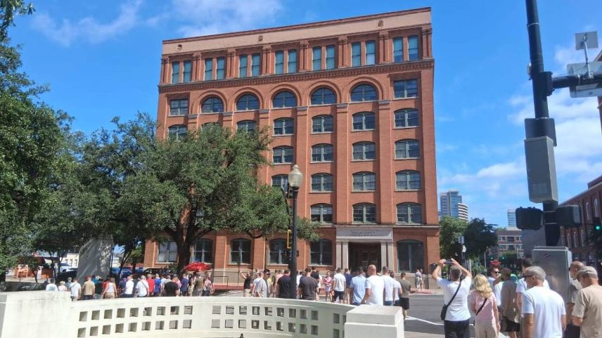Grupo de pessoas caminha em frente ao prédio de tijolos vermelhos do Dallas County Administration Building sob céu azul. Árvore grande e fonte com água parada estão em primeiro plano.