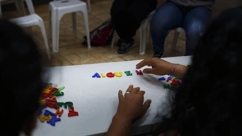 Duas pessoas sentadas em cadeiras brancas manipulam letras plásticas coloridas sobre uma mesa branca. Letras formam parcialmente a palavra 'ALGUEM'. Piso de cerâmica e outras cadeiras ao fundo.