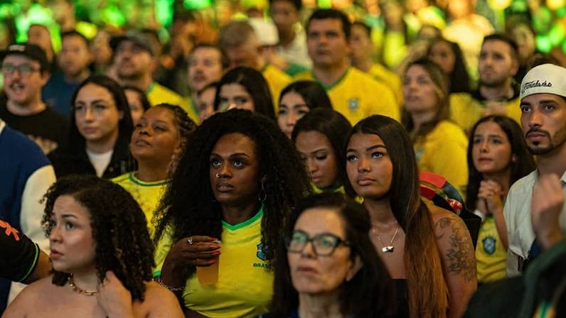 Grupo diversificado de torcedores brasileiros, muitos vestindo camisas amarelas da seleção, observa atentamente um evento. A iluminação verde destaca o ambiente interno, com expressões sérias e concentradas.