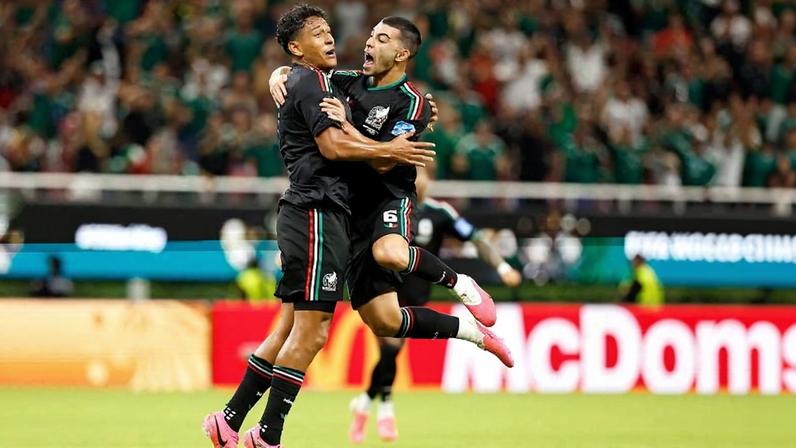 Dois jogadores com uniforme preto e detalhes verdes e vermelhos pulam e se abraçam em campo durante partida. Público ao fundo assiste animado.