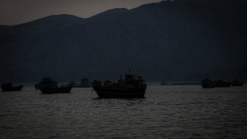 Seis barcos de pesca flutuam em mar calmo sob céu nublado, com montanhas grandes e desfocadas ao fundo.