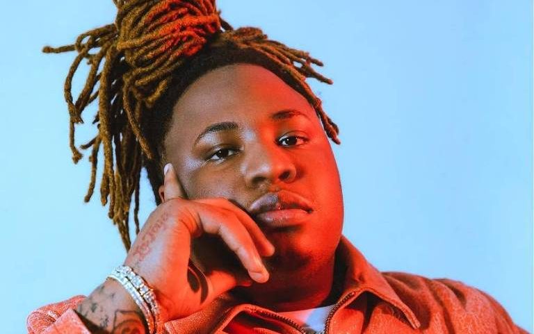 Pessoa com dreadlocks presos em coque alto, usando jaqueta laranja e pulseiras, posa com a mão no rosto contra fundo azul.
