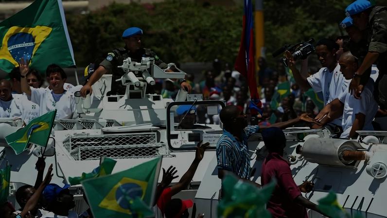 Soldado da ONU em veículo blindado branco segura o troféu da Copa do Mundo enquanto multidão numerosa acena e segura bandeiras do Brasil. Faixa com a palavra 'BIENVENUE' e bandeira brasileira está pendurada acima da rua.