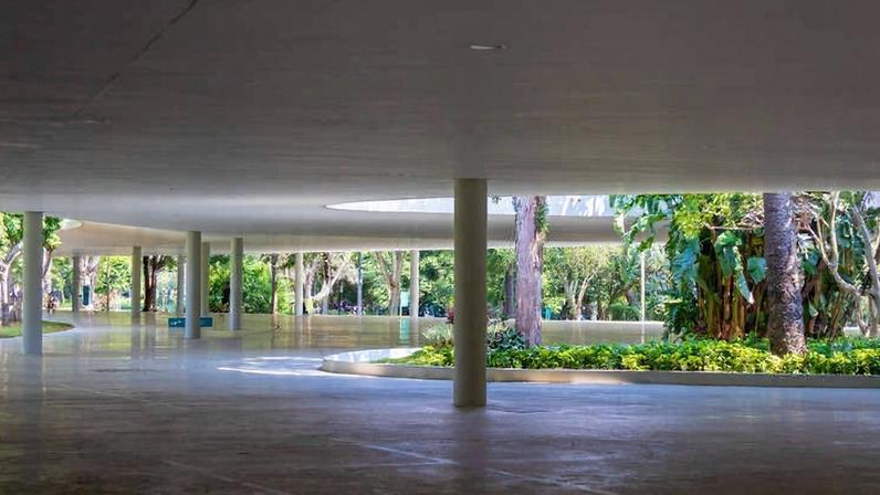Área ampla e coberta com piso liso e teto baixo sustentado por pilares brancos. No centro, há um jardim com árvores e plantas, iluminado pela luz natural que entra por aberturas no teto. Pessoas estão sentadas e caminhando ao redor do jardim, em ambiente que mistura arquitetura moderna e natureza.