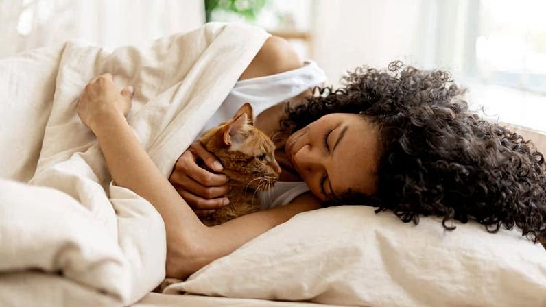 Mulher deitada de lado na cama, abraçando um gato marrom enquanto está coberta por um edredom branco. Luz natural entra pela janela ao fundo.