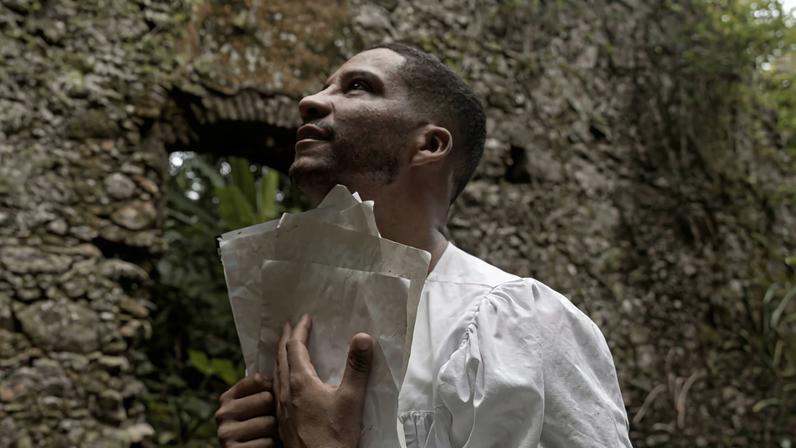 Homem de pele escura veste camisa branca e segura esculturas de pedra enquanto olha para cima. Ele está em frente a uma parede de pedra antiga com uma abertura retangular, cercada por vegetação.
