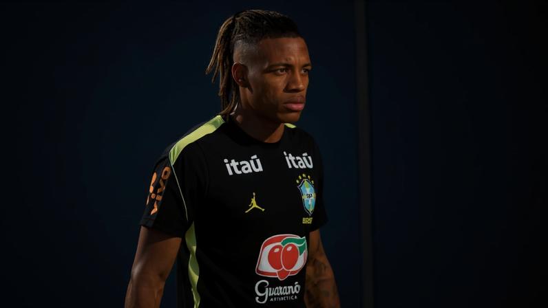 Jogador de futebol brasileiro com cabelo trançado e laterais raspadas veste camisa preta de treino da seleção, com logos de patrocinadores e escudo da CBF, em ambiente externo com rede ao fundo.