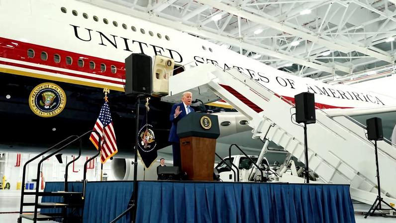 Homem em terno azul fala em púlpito com selo presidencial diante do avião presidencial Air Force One em hangar.