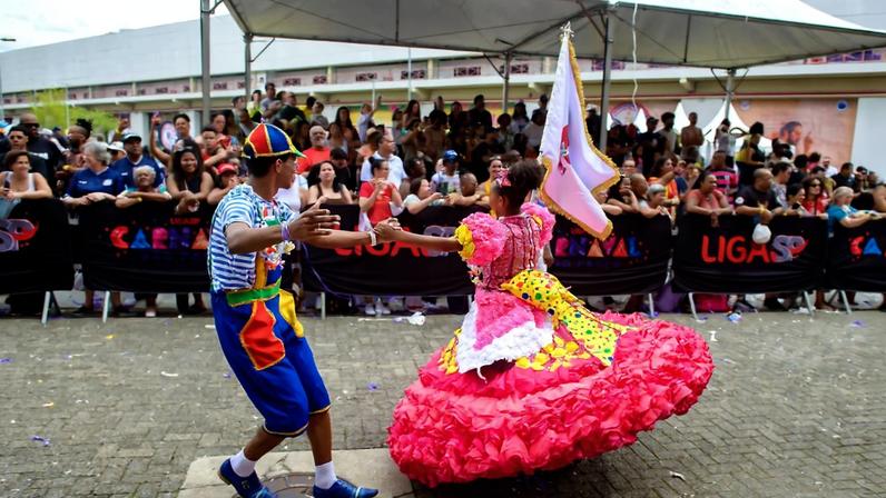 Casal vestido com roupas típicas de mestre-sala e porta-bandeira dança em área externa pavimentada. Mulher usa vestido rosa com babados e segura bandeira, homem veste calça azul, camiseta listrada e chapéu colorido. Público observa atrás de barreira preta com inscrições.