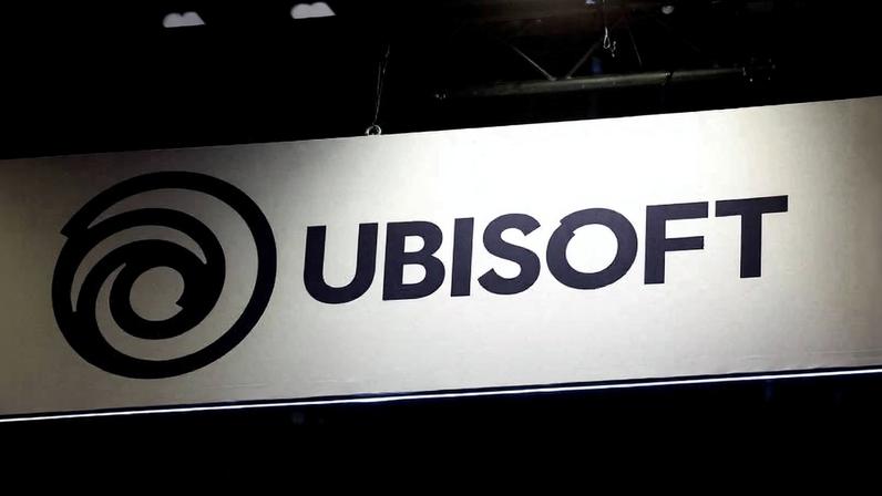 Painel branco com o logotipo preto da Ubisoft, composto por um símbolo circular espiralado à esquerda e a palavra 'UBISOFT' em letras maiúsculas à direita, iluminado em ambiente escuro.