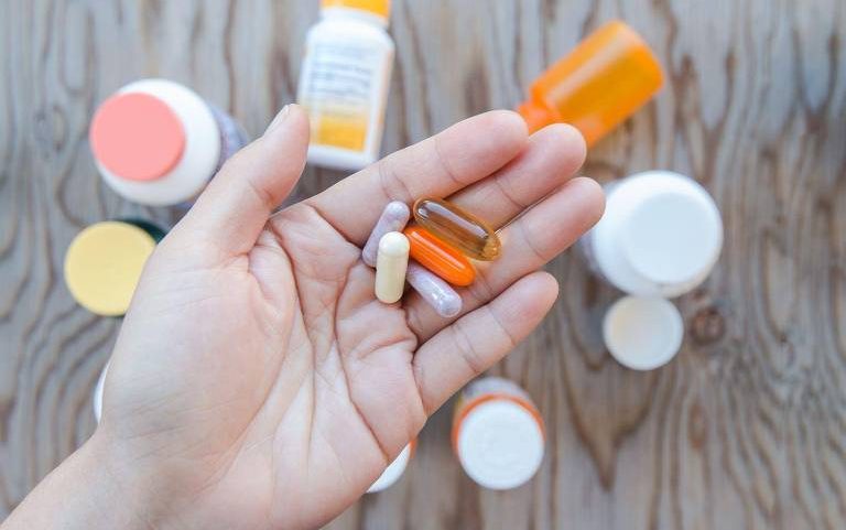 Mão aberta segura cinco cápsulas e comprimidos de cores variadas. Ao fundo, sobre superfície de madeira, há vários frascos de medicamentos desfocados.