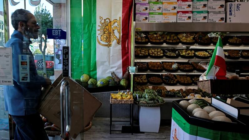 Homem de perfil entra por porta de vidro em mercado decorado com bandeira do Irã. Prateleiras ao fundo exibem frutas variadas organizadas em cestos. Bandeira iraniana também está em cesta próxima à entrada.