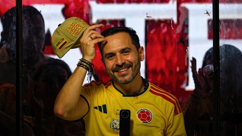 Homem com chapéu e camisa amarela da seleção colombiana segura jornal enquanto crianças e adultos ao redor vestem camisas amarelas da seleção. Ambiente com várias pessoas e câmeras ao fundo.