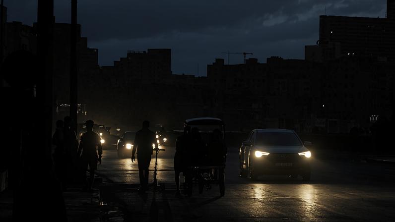 Pessoas caminham e empurram carrinho na rua enquanto carros com faróis acesos passam ao entardecer. Edifícios urbanos ao fundo sob céu com nuvens escuras.