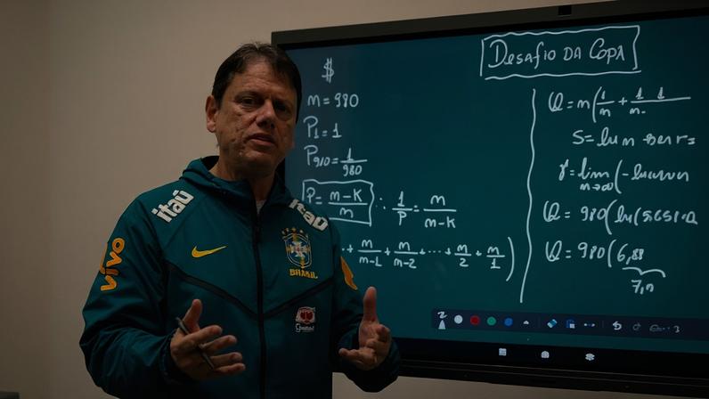 Homem com jaqueta azul da seleção brasileira de futebol está em sala de aula, gesticulando enquanto explica fórmulas matemáticas escritas em quadro digital ao fundo.