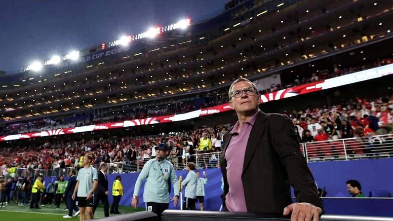 Homem de óculos e blazer escuro apoia a mão em estrutura na frente, com multidão e jogadores ao fundo em estádio iluminado à noite.