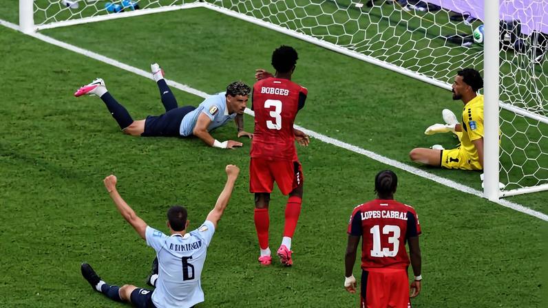 Jogador da seleção do Uruguai sentado no gramado com os braços erguidos em comemoração, enquanto goleiro e dois defensores do time adversário, vestidos de vermelho, estão próximos ao gol. Bola está dentro da rede, indicando gol marcado.