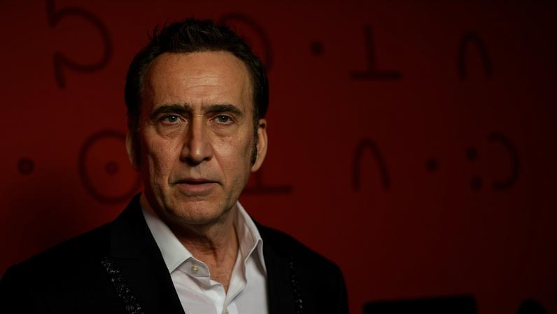 Homem de meia-idade com cabelo curto e escuro veste terno preto e camisa branca, posando diante de fundo vermelho com letras desfocadas.