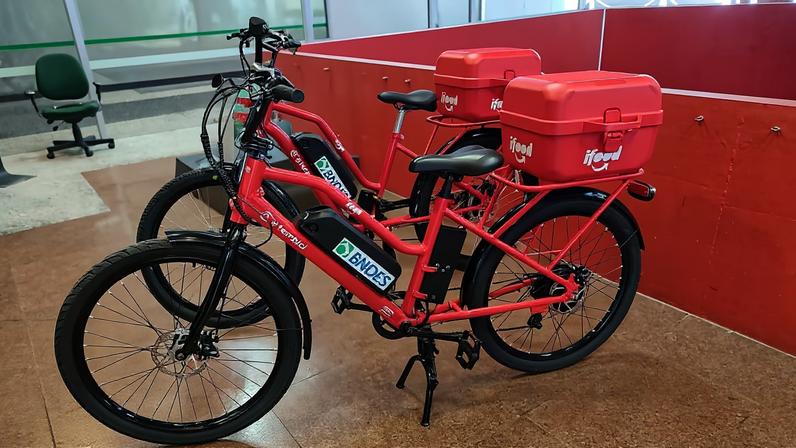 Duas bicicletas vermelhas com caixas térmicas vermelhas fixadas na parte traseira estão estacionadas lado a lado em um piso de cerâmica marrom dentro de um ambiente fechado. As bicicletas têm adesivos com a marca 'BNDES' e 'iFood'. Ao fundo, há divisórias vermelhas e uma cadeira verde próxima a uma mesa.