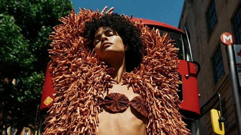 Modelo com casaco e saia texturizados em tons terrosos posa com braços levantados em rua da cidade, com ônibus vermelho ao fundo.