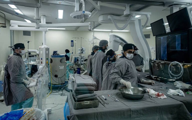 Cinco profissionais de saúde vestindo aventais, toucas e máscaras realizam procedimentos em sala de cirurgia com equipamentos médicos modernos, monitores e instrumentos cirúrgicos organizados em mesa.