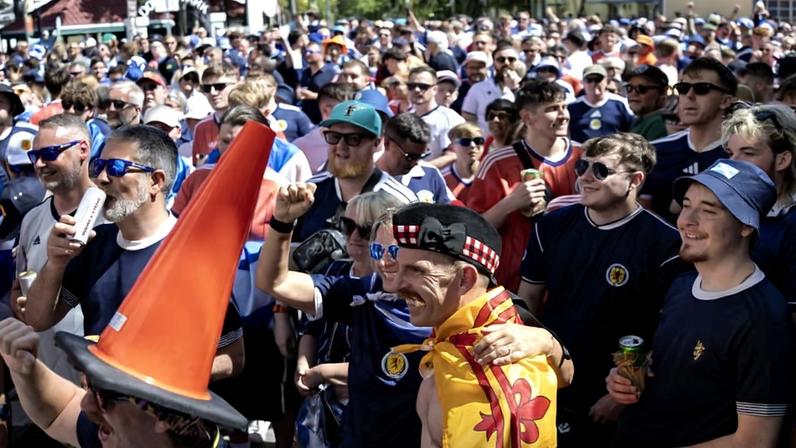 Multidão de torcedores vestindo camisas da seleção da Escócia em evento ao ar livre durante o dia. Um homem usa um chapéu com cone de trânsito e outro está envolto em uma bandeira escocesa com brasão vermelho.