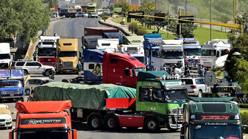 Diversos caminhões de carga ocupam todas as faixas de uma avenida larga em área urbana, causando congestionamento. Ao fundo, há uma grande escultura metálica de figura humana e prédios residenciais visíveis.