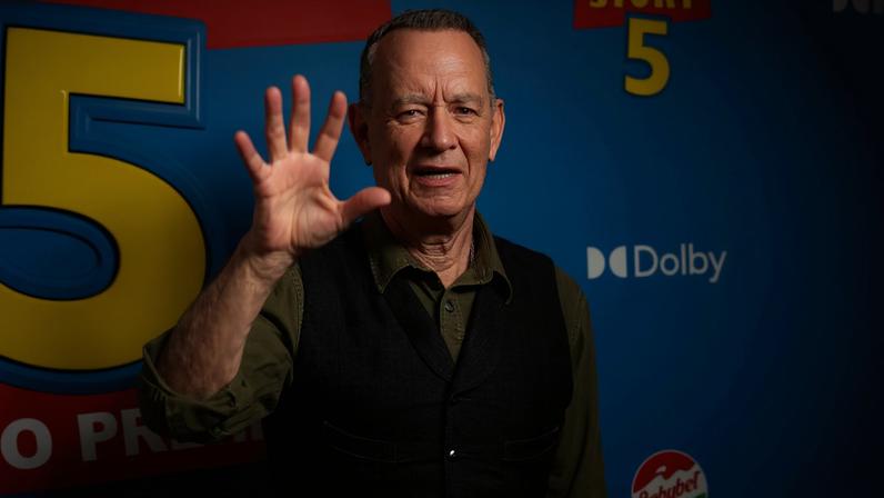 Homem de meia-idade com colete preto sobre camisa verde-oliva levanta a mão direita em gesto de saudação diante de fundo azul com logo de Toy Story 5 e marcas Dolby e Babybel.