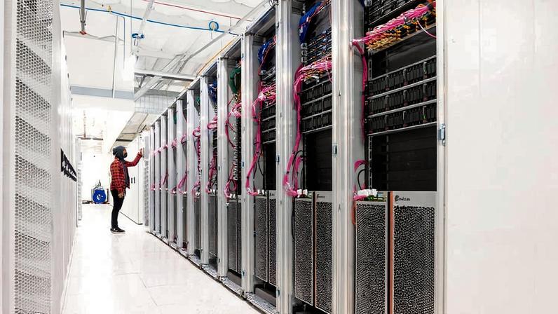 Pessoa com camisa xadrez vermelha e preta verifica equipamentos em uma fileira de racks de servidores em sala branca e iluminada de data center.