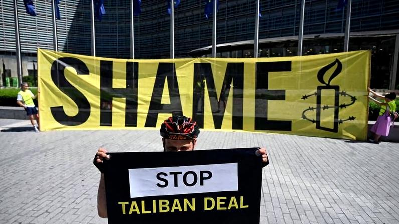 Pessoa com capacete de bicicleta segura cartaz preto com texto "STOP TALIBAN DEAL" em frente a faixa amarela grande com a palavra "SHAME" e o logo da Anistia Internacional, diante de edifício moderno com bandeiras azuis.