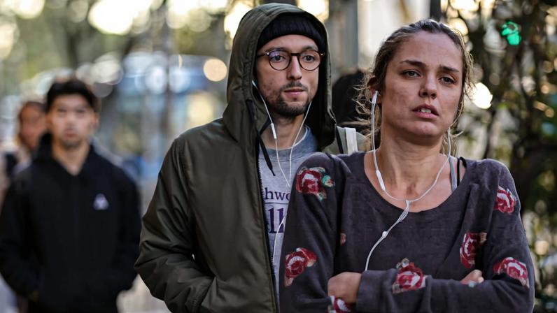 Homem de jaqueta verde com capuz e óculos caminha atrás de mulher com blusa cinza estampada, ambos usando fones de ouvido. Outras pessoas aparecem desfocadas ao fundo em ambiente externo.