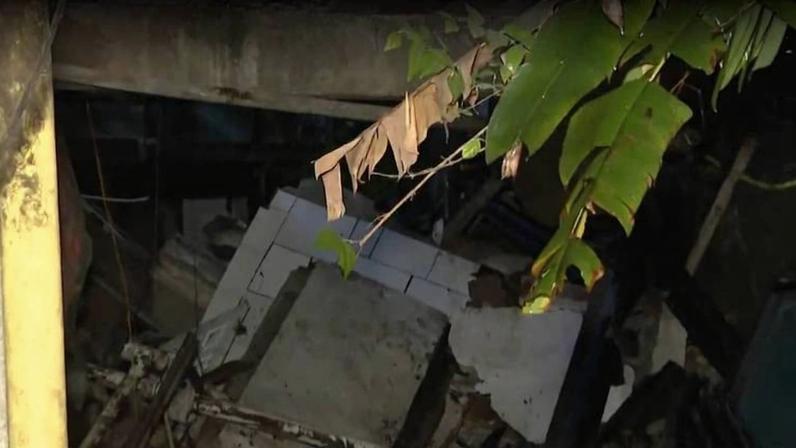 Sobrado parcialmente destruído com escombros e paredes caídas em área escura, com vegetação visível no canto superior direito. Cena iluminada artificialmente, indicando ocorrência noturna.