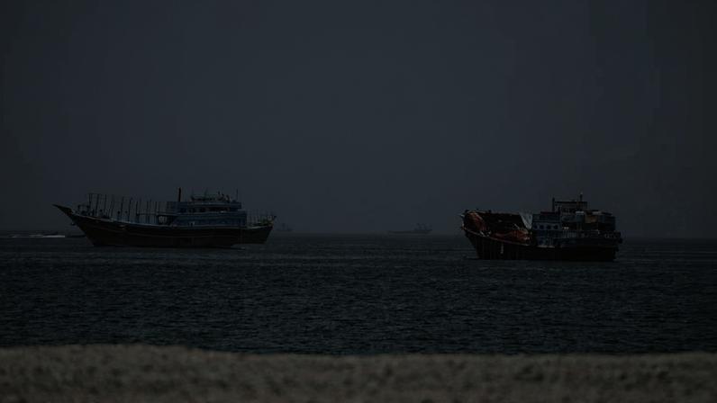 Dois barcos de madeira pintados de azul navegam em mar calmo sob céu nublado. A praia de areia clara aparece em primeiro plano.