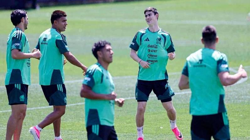 Cinco jogadores de futebol com coletes verdes e uniformes escuros realizam exercícios em campo gramado durante treino ao ar livre em dia ensolarado.