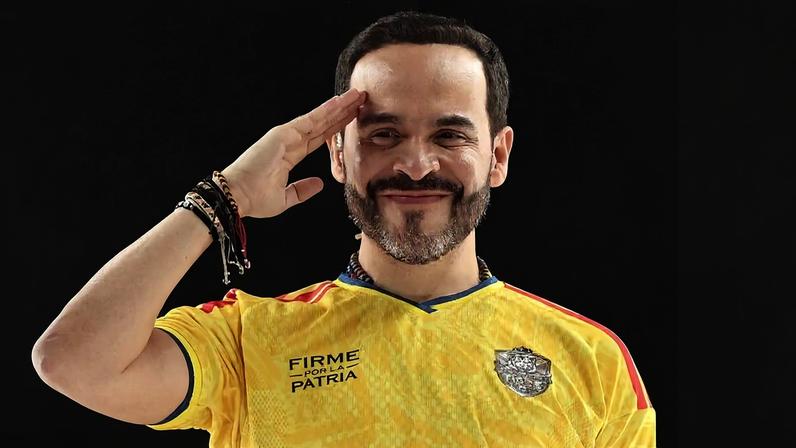 Homem de cabelo curto e barba faz saudação com a mão direita na testa. Ele veste camiseta amarela com detalhes vermelhos e escudo no peito, com a frase 'FIRME PELA PÁTRIA'. Fundo escuro.