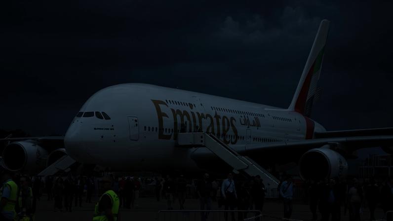 Avião Airbus A380 da Emirates estacionado em pista com escadas de embarque acopladas. Pessoas caminham ao redor e observam a aeronave sob céu nublado.