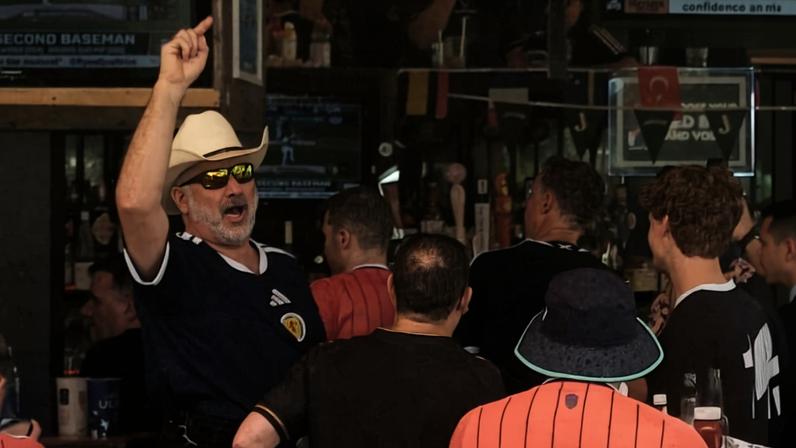 Grupo de torcedores vestidos com camisas listradas e kilts assiste a jogo de futebol em bar. Vários televisores exibem a partida, e um guarda-sol central divide o ambiente interno e externo.