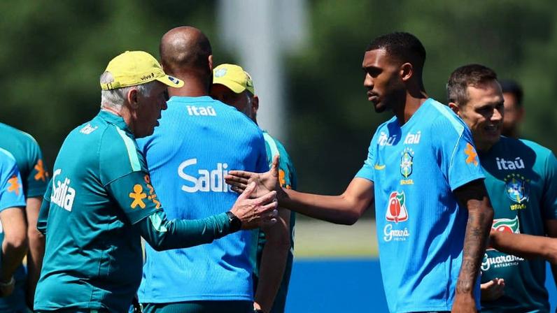Membros da seleção brasileira de futebol se cumprimentam durante treino ao ar livre em dia ensolarado. Homem mais velho com boné amarelo e uniforme verde cumprimenta jogador jovem de camiseta azul com símbolos da CBF e patrocinadores.