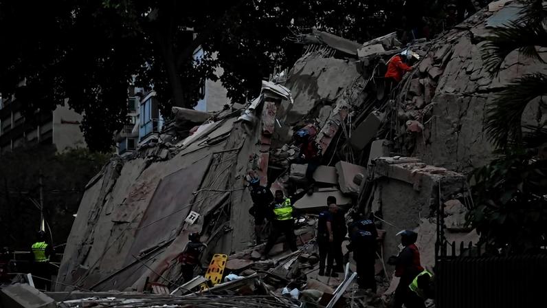 Equipes de resgate trabalham entre escombros de prédio desabado em área urbana. Pessoas usam capacetes e coletes, escalam destroços e auxiliam na busca por sobreviventes.
