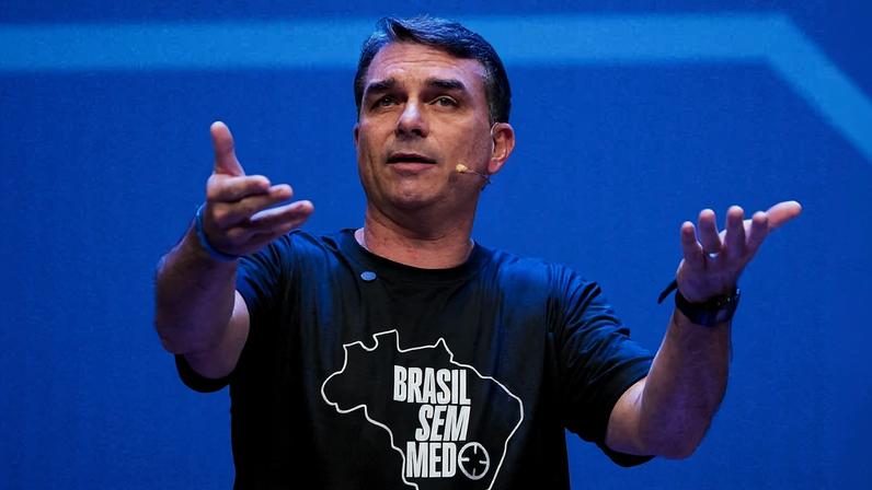 Homem de meia-idade com camiseta preta estampada com mapa do Brasil e texto 'Brasil Sem Medo' gesticula com as mãos abertas em palco com fundo azul.